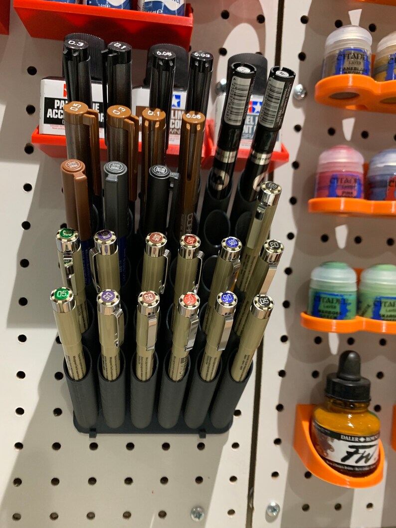 May include: A black plastic pen holder with 18 individual slots for pens and markers. The holder is mounted on a white pegboard wall. The holder is filled with a variety of pens and markers, including black, brown, and gold colored pens.