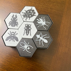 May include: A hexagonal game board with six black and white tiles. Each tile features a different insect illustration, including a ladybug, spider, ant, bee, beetle, and a moth.