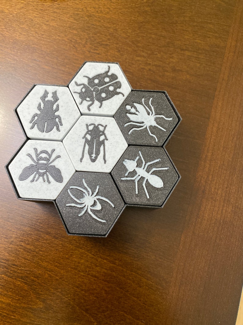 May include: A hexagonal game board with six black and white tiles. Each tile features a different insect illustration, including a ladybug, spider, ant, bee, beetle, and a moth.
