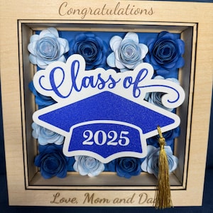 May include: A wooden shadow box with a graduation theme. The box features paper flowers in shades of blue. The words "Congratulations", "Class of 2025", and "Love, Mom and Dad" are visible. A blue graduation cap with a gold tassel is also included.