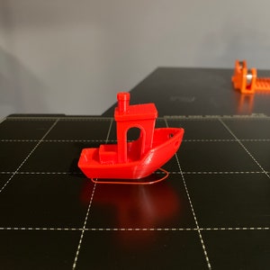 May include: A small, red, 3D printed toy boat with a chimney on the top. The boat is sitting on a black surface.