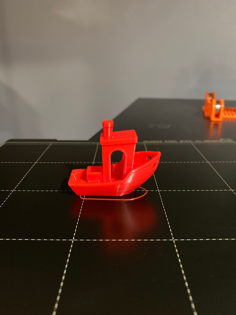 May include: A small, red, 3D printed toy boat with a chimney on the top. The boat is sitting on a black surface.