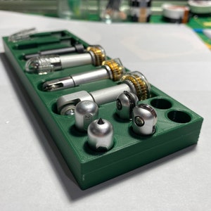 May include: A green plastic storage case with various silver and gold metal tools. The tools are arranged in the case with the tips facing up. The case has a number "21" printed on it.