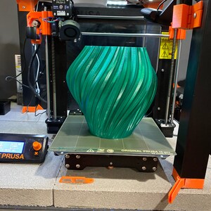 May include: A green 3D printed vase with a swirling pattern is on the print bed of a black 3D printer. The printer is labeled "ORIGINAL PRUSA i3 MK3" and has a black and orange frame.