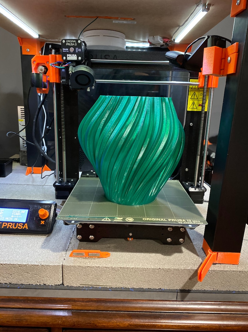 May include: A green 3D printed vase with a swirling pattern is on the print bed of a black 3D printer. The printer is labeled "ORIGINAL PRUSA i3 MK3" and has a black and orange frame.