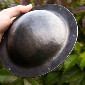 May include: A round, black metal shield with a hammered surface and a slightly raised rim. The shield has several small holes around the edge.