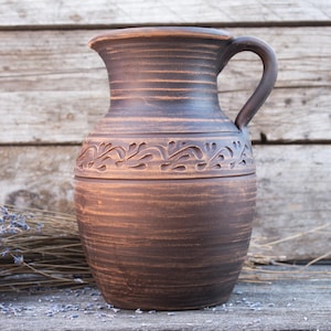 Könnte beinhalten: Ein brauner Keramikkrug mit Henkel, mit einem dekorativen Blumenband in der Mitte. Der Krug hat ein rustikales, handwerkliches Aussehen und steht vor einem hölzernen Hintergrund. Getrockneter Lavendel ist im Vordergrund.