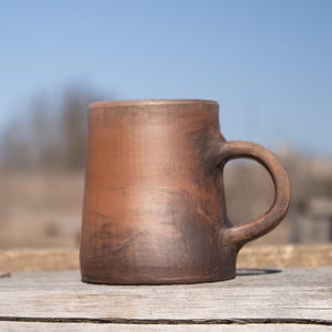 Peut inclure: Une tasse en céramique marron faite à la main avec une anse, sur un fond extérieur flou. La tasse a un aspect rustique, avec des variations de couleur et de texture. La tasse est posée sur une surface en bois.