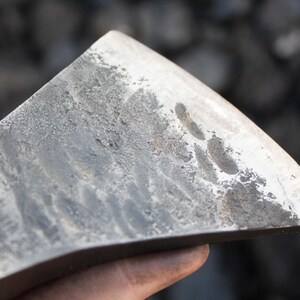 Pode incluir: Close-up de uma cabe&ccedil;a de machado de metal cinza desgastada com uma l&acirc;mina ligeiramente curva. A superf&iacute;cie da cabe&ccedil;a do machado mostra sinais de uso e desgaste.