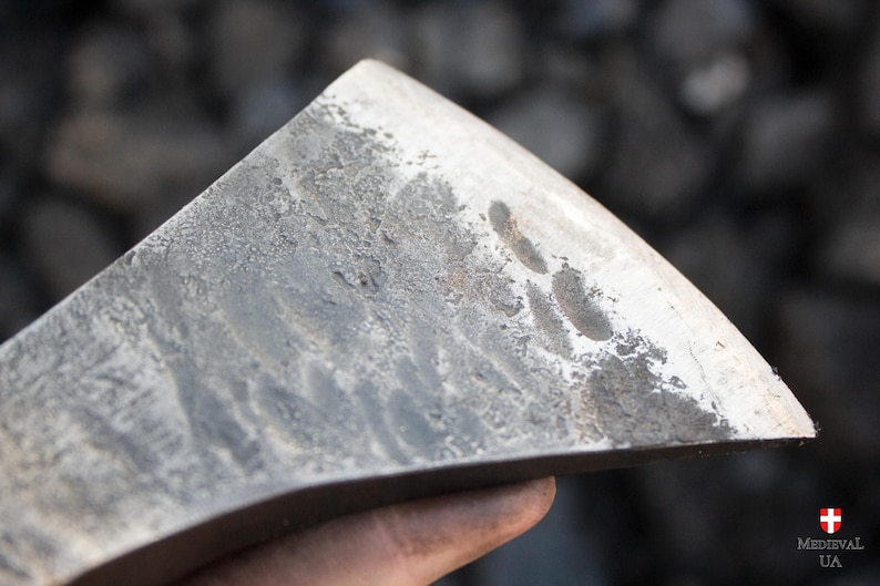 Pode incluir: Close-up de uma cabe&ccedil;a de machado de metal cinza desgastada com uma l&acirc;mina ligeiramente curva. A superf&iacute;cie da cabe&ccedil;a do machado mostra sinais de uso e desgaste.