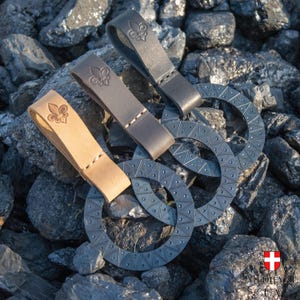 Puede incluir: Tres lazos de cinturón de cuero con un diseño de flor de lis. Los lazos están unidos a tres anillos de metal negro con un patrón decorativo.