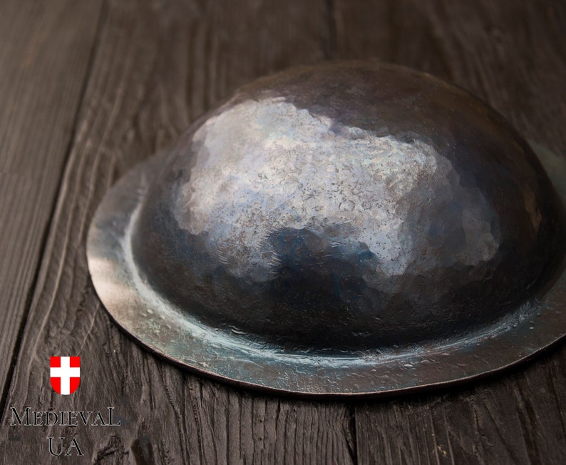 Puede incluir: Un escudo redondo de metal martillado con un borde ligeramente elevado. El escudo es de color gris oscuro con un brillo met&aacute;lico.