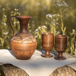 May include: A brown clay pitcher with a curved handle and decorative carved patterns around the neck, accompanied by two matching brown clay goblets with stems. The set is placed on a light-colored fabric cloth.