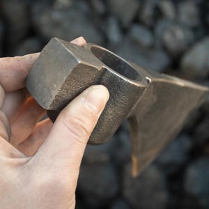 Pode incluir: Uma m&atilde;o segura uma cabe&ccedil;a de machado de metal com um cabo quadrado. A cabe&ccedil;a do machado &eacute; feita de a&ccedil;o e tem uma l&acirc;mina afiada. O machado &eacute; uma ferramenta tradicional usada para cortar madeira.