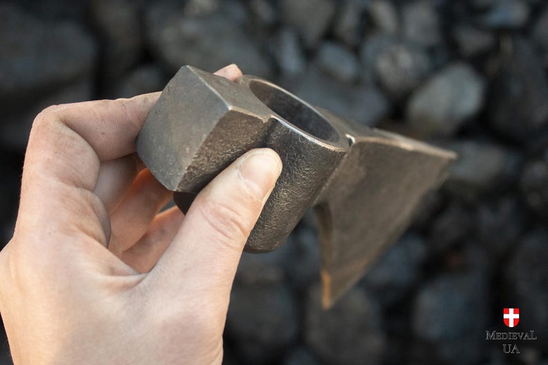 Pode incluir: Uma m&atilde;o segura uma cabe&ccedil;a de machado de metal com um cabo quadrado. A cabe&ccedil;a do machado &eacute; feita de a&ccedil;o e tem uma l&acirc;mina afiada. O machado &eacute; uma ferramenta tradicional usada para cortar madeira.