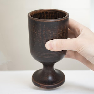 May include: A dark brown, handmade ceramic goblet with a wide rim and a pedestal base. The goblet is held by a hand, showcasing its rustic texture and design. The goblet is approximately 5 inches tall.