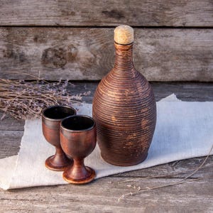 Könnte beinhalten: Eine braune Keramikflasche mit gerippter Textur und hellem Korkverschluss, begleitet von zwei passenden Keramikbechern. Das Set weist eine dunkle, verwitterte Oberfläche mit sichtbaren horizontalen Rillen auf dem Flaschenkörper auf.
