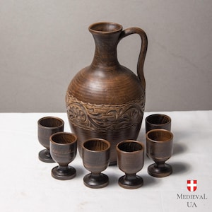 May include: A rustic brown ceramic pitcher with a handle and carved floral design, accompanied by six small matching wooden cups. The set is arranged on a white surface, suggesting a traditional or vintage aesthetic.