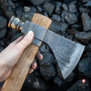 May include: A hand holds a  tomahawk with a wooden handle and a metal head. The head is a dark gray color and has a slight curve to it. The handle is a light brown color and has a slight grain to it. The tomahawk is a traditional weapon used by Native Americans.