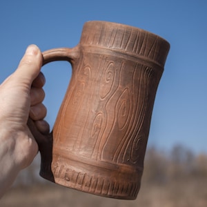Puede incluir: Una jarra de cerámica marrón con asa, con un diseño de vetas de madera. La jarra se sostiene contra un fondo de cielo azul. La textura y el color de la jarra sugieren una estética rústica y artesanal, adecuada para bebidas.