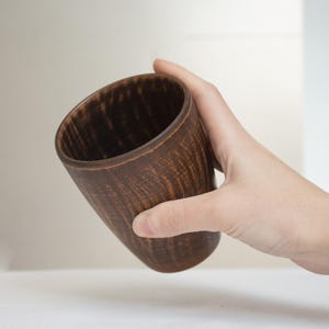Peut inclure: Une tasse en bois marron faite à la main avec une surface texturée. La tasse est tenue en biais, mettant en valeur son intérieur et le grain naturel du bois. La tasse a une esthétique rustique et médiévale.