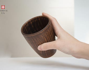 May include: A handcrafted, brown wooden cup with a textured surface. The cup is held at an angle, showcasing its interior and the natural wood grain. The cup has a rustic, medieval aesthetic.