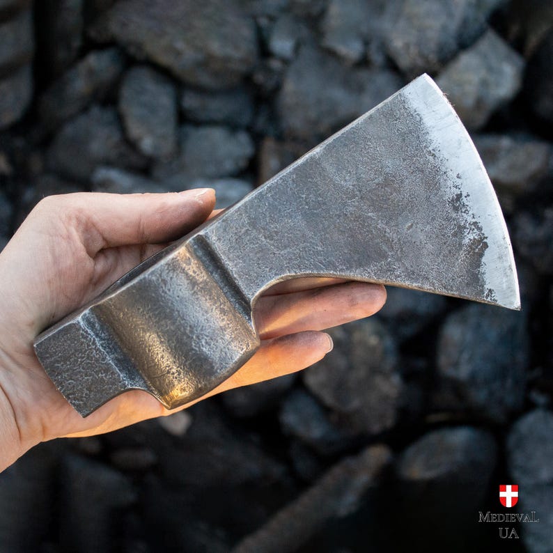 Pode incluir: Uma m&atilde;o segura um machado de metal cinza grande, com uma l&acirc;mina ligeiramente curva. O machado tem uma superf&iacute;cie texturizada e um cabo arredondado.