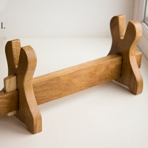 May include: A wooden sword stand with two upright supports and a horizontal bar. The stand is made of light brown wood and has a simple design.