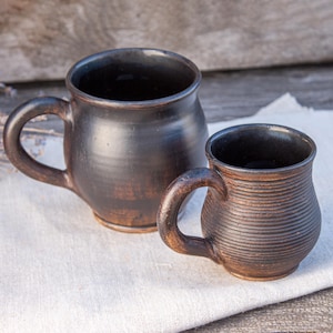 May include: Two handmade, dark brown ceramic mugs with handles. The larger mug has a smooth surface, while the smaller one features horizontal ridges. The mugs are set on a white cloth against a rustic wooden background.