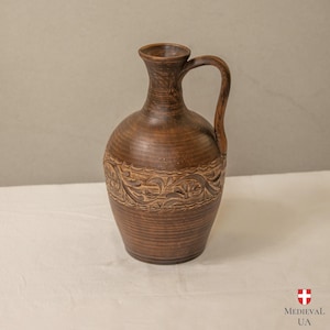 Puede incluir: Una jarra de cerámica marrón con asa. La jarra tiene un cuerpo redondeado y un cuello estrecho. Un patrón floral decorativo está tallado alrededor del centro. La jarra está sobre una superficie blanca. Cerámica artesanal.