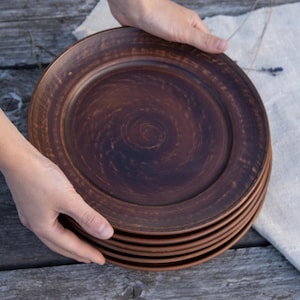 Puede incluir: Una pila de platos redondos de cerámica de color marrón oscuro con un acabado liso y ligeramente moteado, con un borde ancho. Los platos presentan un sutil patrón de remolinos circulares en la superficie, con tonos naturales y terrosos.