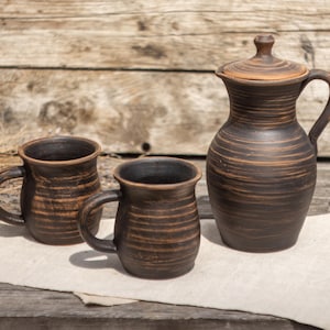 May include: A set of two dark brown clay mugs and a matching lidded pitcher featuring horizontal ridged textures. The pottery has a rustic, unglazed appearance with visible wheel-thrown markings and a deep, earthy colour palette throughout the pieces.