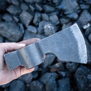 May include: A hand holds a forged steel axe head with a textured, gray finish. The axe head has a curved blade and a rectangular handle socket. The background is a pile of dark gray rocks.