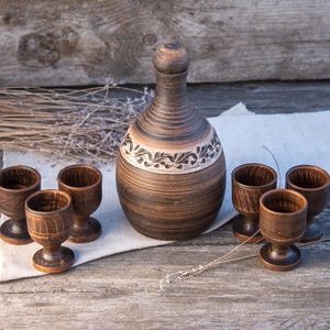 May include: A brown clay decanter with a rounded body, a narrow neck, and a matching stopper, featuring a carved leaf pattern around the center. The set includes four small, matching brown clay stemmed cups with a textured, dark finish.