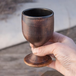 May include: A handcrafted, dark brown wooden goblet with a wide base and a rounded bowl. The goblet is held in a hand, showcasing its smooth, polished surface. The goblet is approximately 13 cm tall.