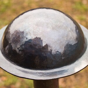 Puede incluir: Primer plano de un casco redondo de plata con una parte superior abovedada. El casco est&aacute; hecho de metal y tiene un acabado liso y pulido.