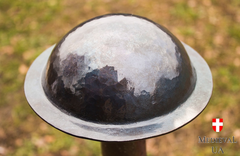 Puede incluir: Primer plano de un casco redondo de plata con una parte superior abovedada. El casco est&aacute; hecho de metal y tiene un acabado liso y pulido.