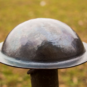 Puede incluir: Un casco de metal martillado con una parte superior redondeada y un borde ancho. El casco es de color gris oscuro y tiene una superficie texturizada.
