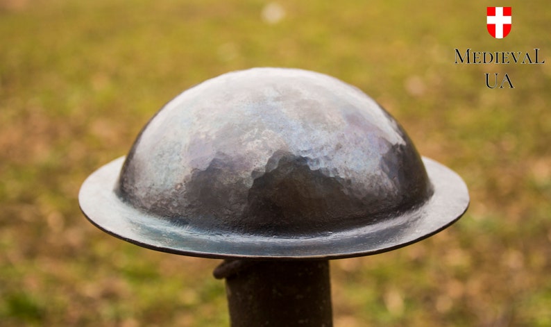 Puede incluir: Un casco de metal martillado con una parte superior redondeada y un borde ancho. El casco es de color gris oscuro y tiene una superficie texturizada.