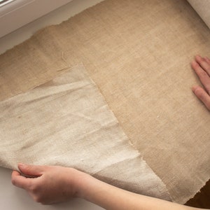 May include: A large roll of natural linen fabric is being unrolled on a white surface. The fabric is a light beige color and has a rough texture. The fabric is being held by a hand.