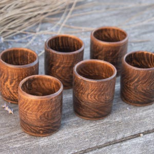 May include: Six handcrafted wooden cups with a rich, dark brown finish. The cups have a cylindrical shape and a visible wood grain pattern. The cups are arranged on a weathered wooden surface, suggesting a rustic or natural theme.