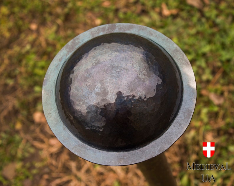 Puede incluir: Un escudo redondo de metal con un acabado martillado. El escudo es de color gris oscuro con un borde azul claro. El escudo est&aacute; en un palo de madera.