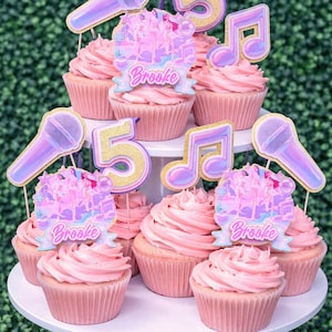 May include: Pink frosted cupcakes on a white tiered stand, decorated with music-themed toppers. Toppers include microphones, musical notes, the number 5, and banners that read "Brooke". The background is a green hedge.