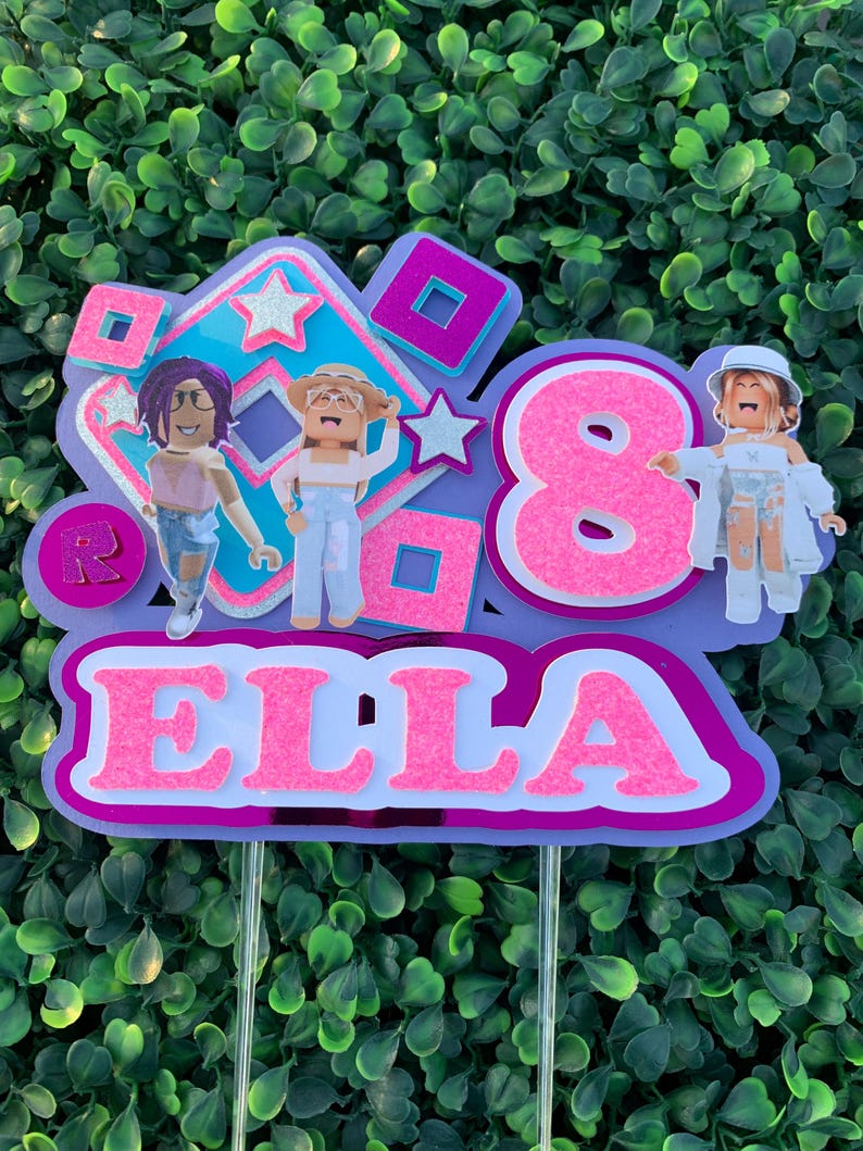 Peut inclure: Un topper de g&acirc;teau rose et violet avec le chiffre 8, le nom Ella et trois personnages Roblox. Les personnages portent des v&ecirc;tements roses, bleus et blancs.