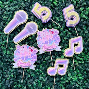 May include: A collection of party decorations, including two microphones, two musical notes, two number 5s, and two banner-shaped toppers with the name "Brooke". The decorations are in shades of purple, pink, and gold, with glitter accents.