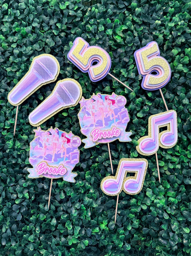 May include: A collection of party decorations, including two microphones, two musical notes, two number 5s, and two banner-shaped toppers with the name "Brooke". The decorations are in shades of purple, pink, and gold, with glitter accents.