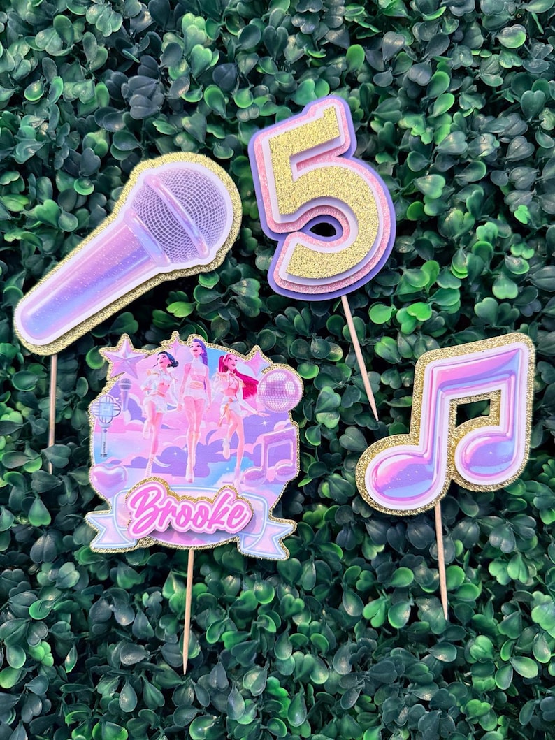 May include: A collection of glittery cake toppers, including a microphone, a musical note, the number 5, and a banner that says "Brooke". The toppers are in shades of pink, purple, and gold, with wooden sticks.