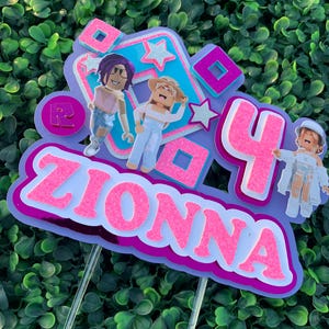 Peut inclure: Un topper de g&acirc;teau rose et violet avec le nom "ZIONNA" et le chiffre "4" dessus. Il y a deux personnages de dessin anim&eacute; sur le topper, l'un avec des cheveux violets et l'autre avec des cheveux blonds. Le topper est d&eacute;cor&eacute; d'&eacute;toiles et de carr&eacute;s.