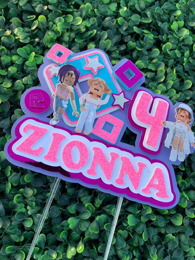 Peut inclure: Un topper de g&acirc;teau rose et violet avec le nom "ZIONNA" et le chiffre "4" dessus. Il y a deux personnages de dessin anim&eacute; sur le topper, l'un avec des cheveux violets et l'autre avec des cheveux blonds. Le topper est d&eacute;cor&eacute; d'&eacute;toiles et de carr&eacute;s.
