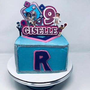 Peut inclure: Un g&acirc;teau carr&eacute; bleu et rose avec un '9' rose et blanc et le nom 'Giselle' sur le dessus. Le g&acirc;teau est d&eacute;cor&eacute; de paillettes argent&eacute;es et a un 'R' rose et violet sur le devant.